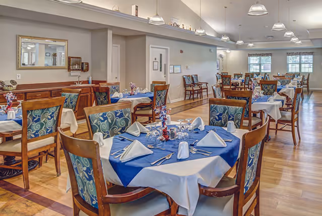 Bright dining room with multiple round tables set with blue and white linens and wooden chairs under pendant lights.
