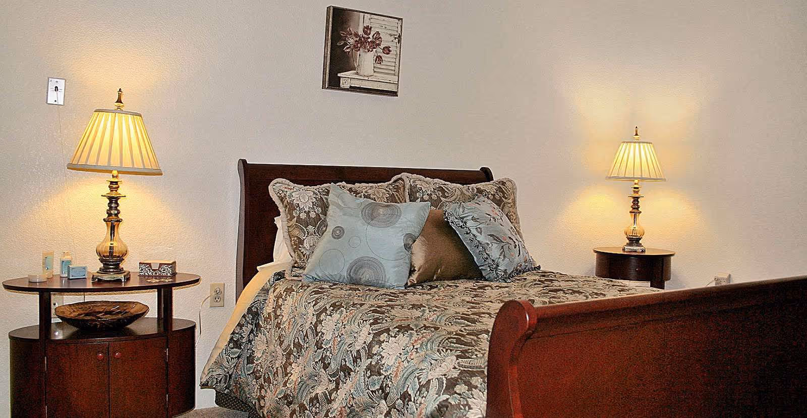 A cozy bedroom with a wooden sleigh bed adorned with multiple decorative pillows and a floral patterned bedspread. There are two bedside tables, each with a lamp emitting warm light. A framed picture of flowers hangs on the wall above the bed.