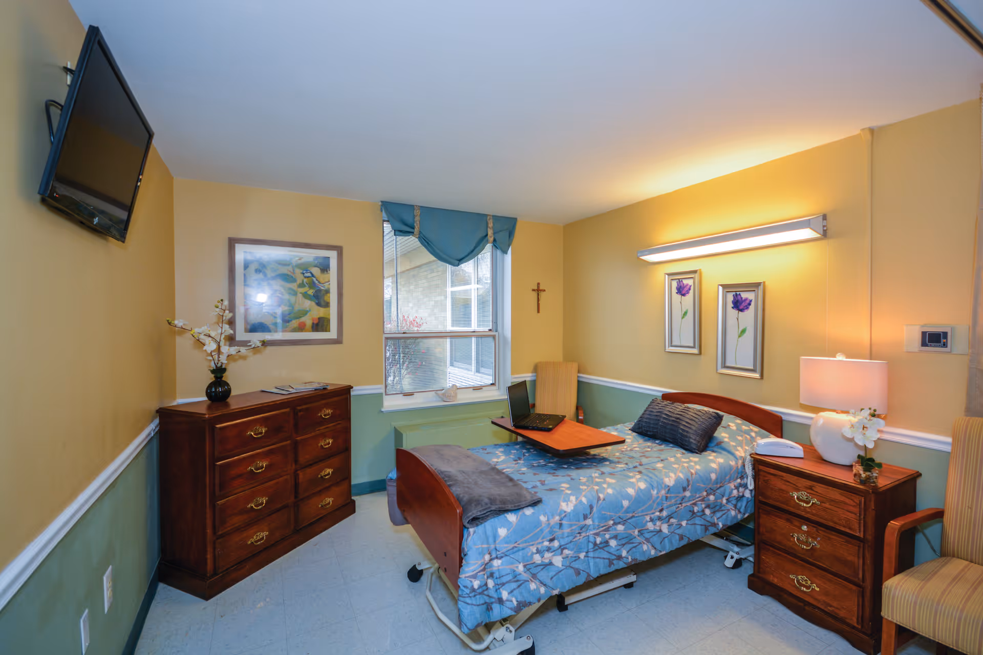 A furnished senior care bedroom with a single bed, bedside tables, dresser, wall-mounted TV and a window.