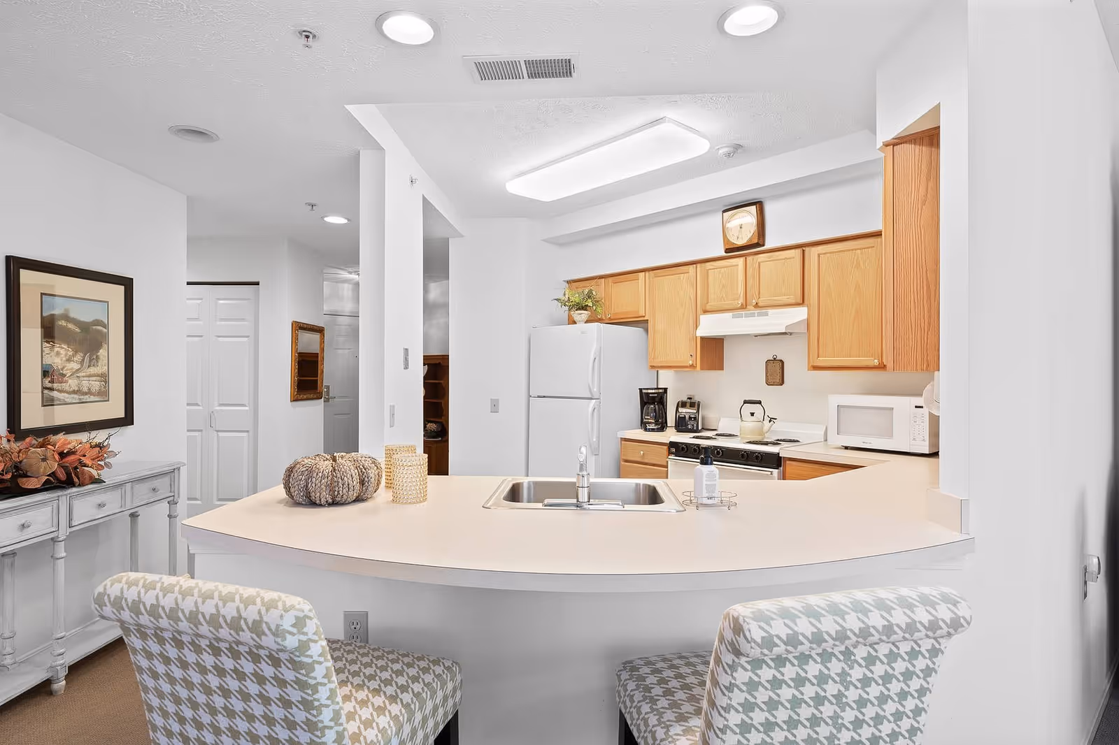 A bright kitchen area with light wood cabinets, a white refrigerator, stove, microwave, and coffee maker. A curved countertop with a built-in sink extends into the foreground with two patterned upholstered chairs placed at the counter. The walls are white, and there is a framed picture and a small console table with decorative items on the left side.