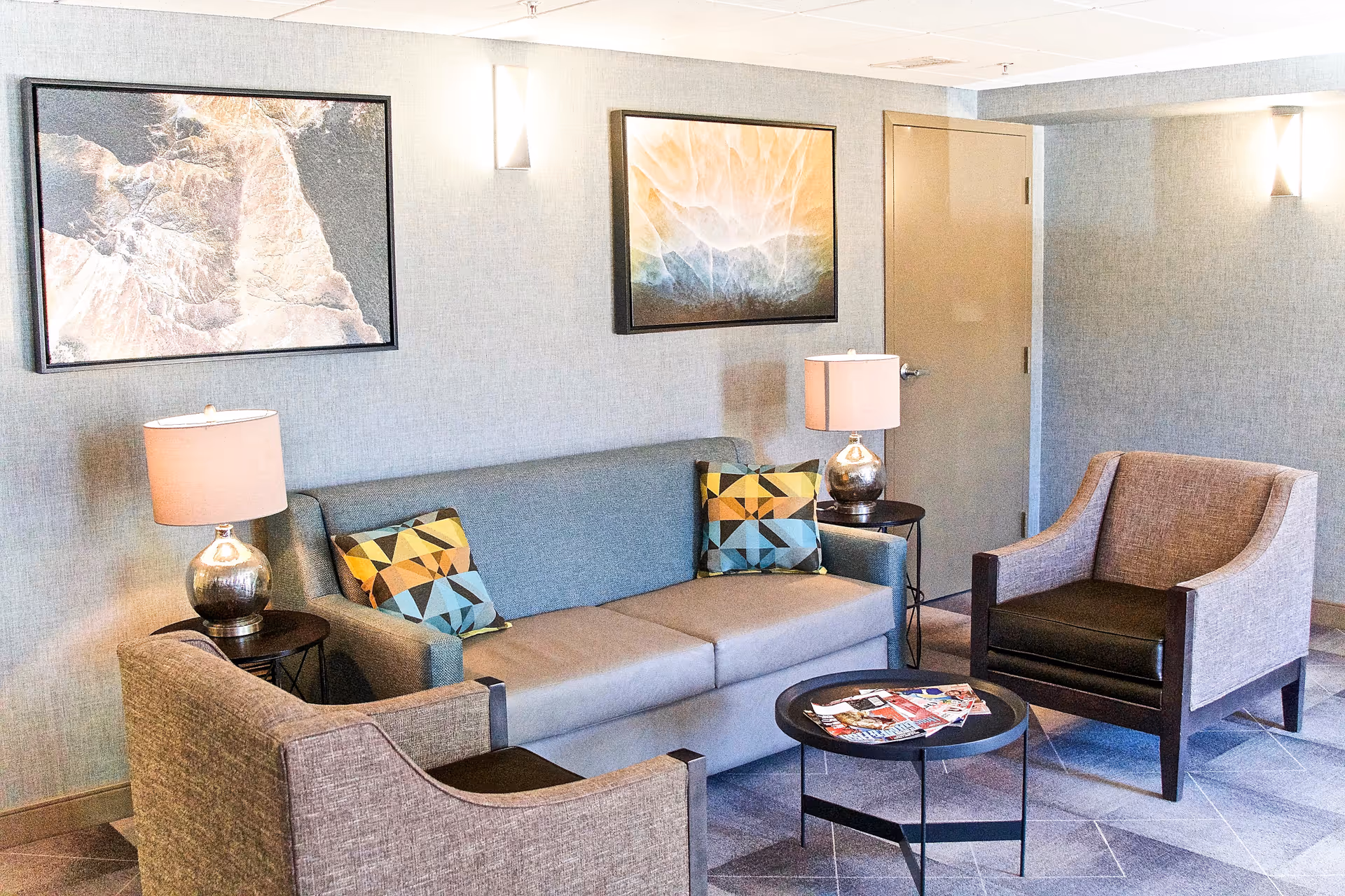 A cozy living room area with a gray sofa adorned with two colorful geometric patterned pillows, two matching armchairs, two side tables each with a lamp, a round black coffee table with magazines on it, and two abstract paintings on the wall behind the sofa.