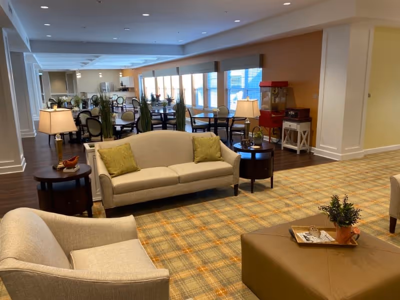 Bright communal living and dining area with sofas, armchairs, tables, lamps, and large windows.