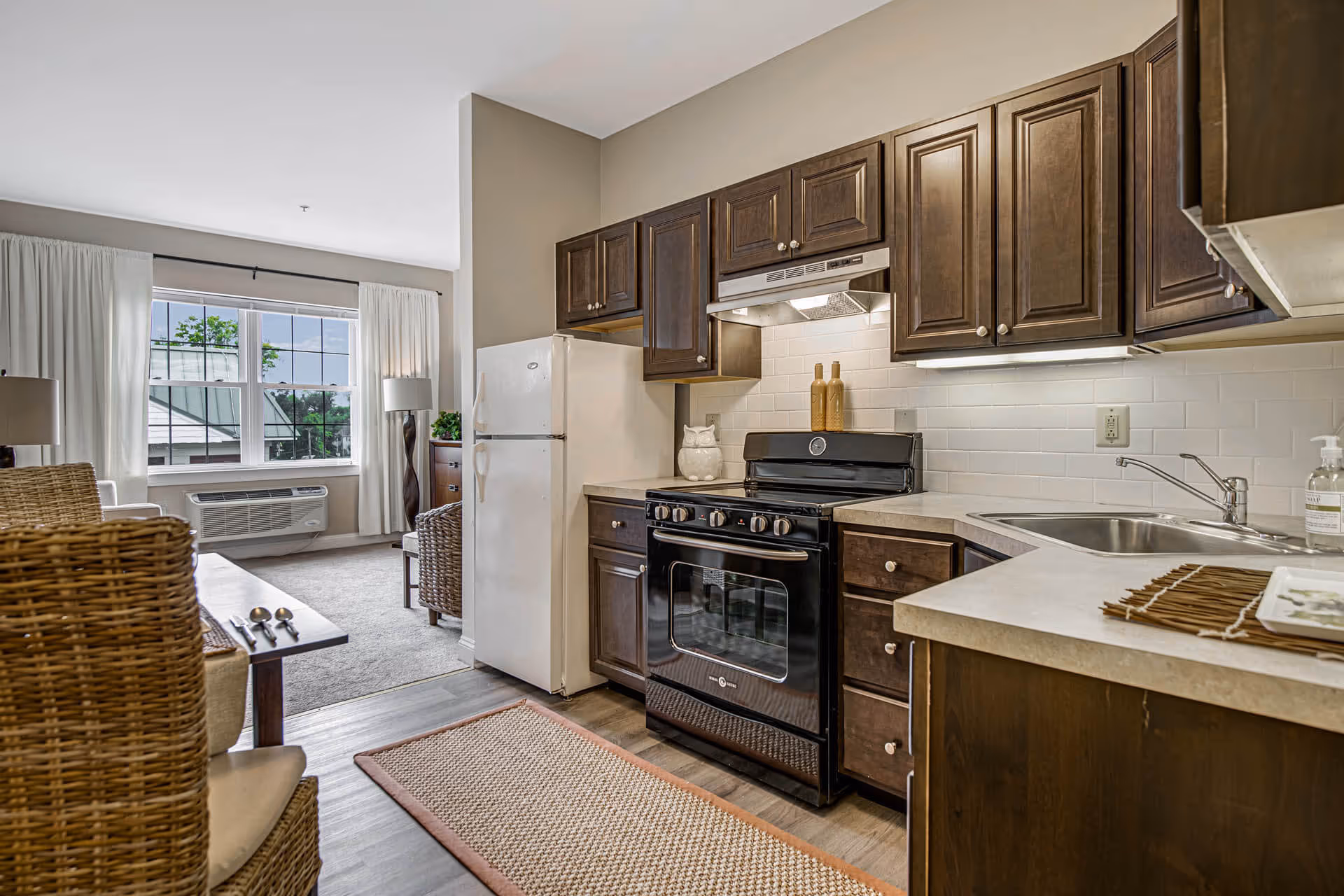 A kitchen area with dark wooden cabinets, a black stove with two decorative bottles on top, a white refrigerator, and a sink with a soap dispenser. The kitchen opens into a living area with a large window, white curtains, a floor lamp, and wicker chairs around a dining table.
