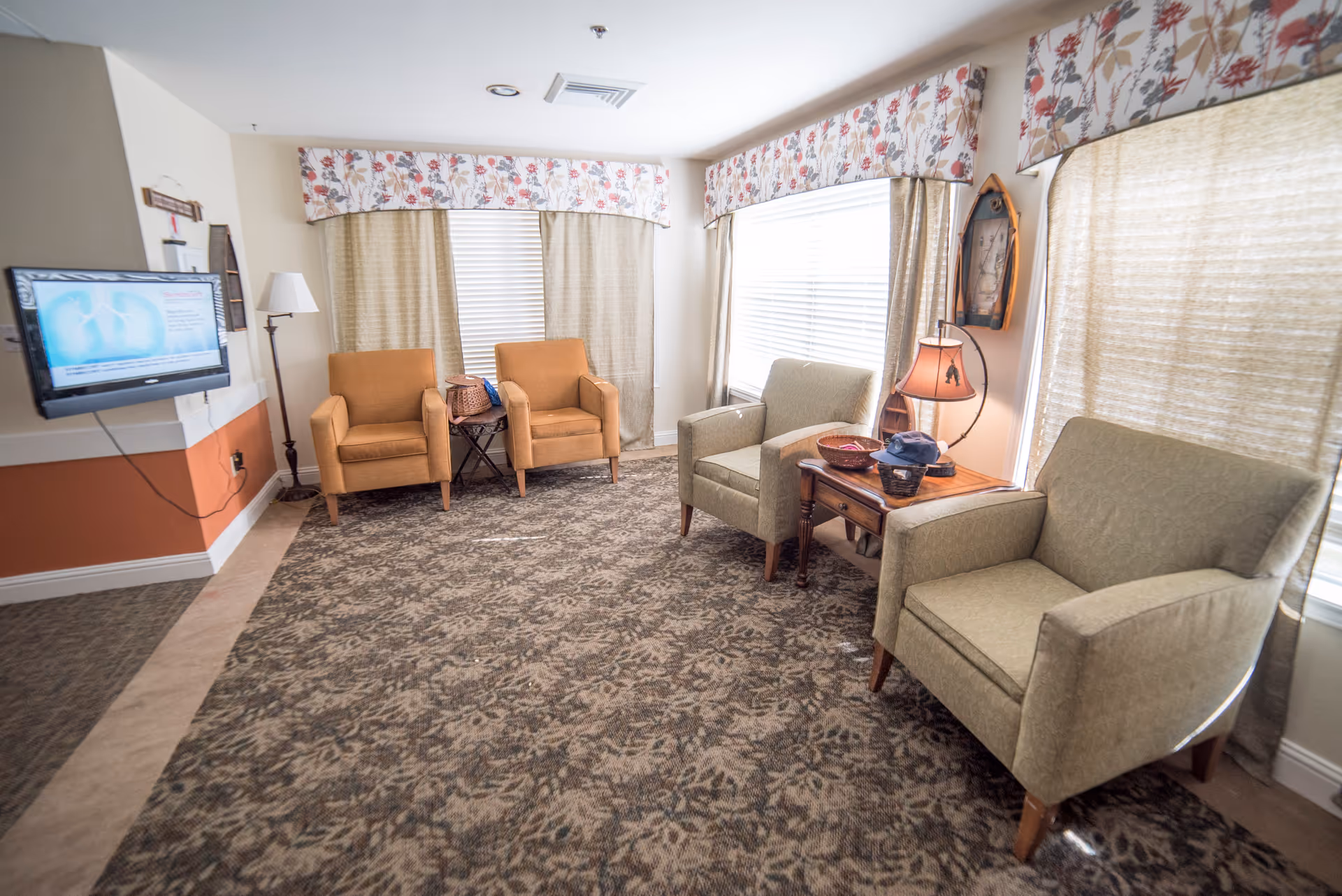 A cozy living room area with four armchairs arranged around a small wooden side table with a lamp and decorative items. The room has patterned carpet, floral valances over the windows, and a wall-mounted TV displaying a medical image.