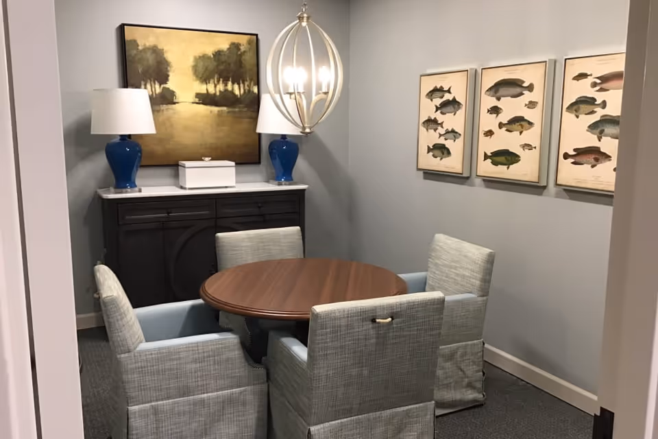 A small dining room with a round wooden table surrounded by four upholstered chairs. On the back wall, there is a dark wooden sideboard with a white top, two blue table lamps with white shades, and a landscape painting above it. On the right wall, there are three framed pictures of various fish species. A modern chandelier with exposed bulbs hangs from the ceiling.