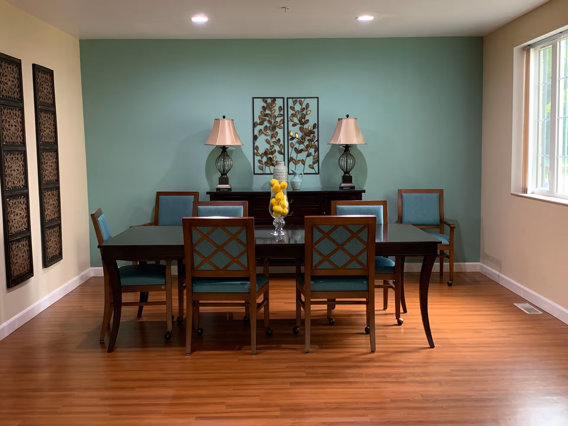 A dining room with a wooden table surrounded by six chairs with teal cushions. On the table is a glass vase filled with yellow lemons. Against the teal accent wall is a dark wooden sideboard with two matching lamps and decorative wall art featuring metal leaves. The room has wooden flooring and a large window on the right letting in natural light.