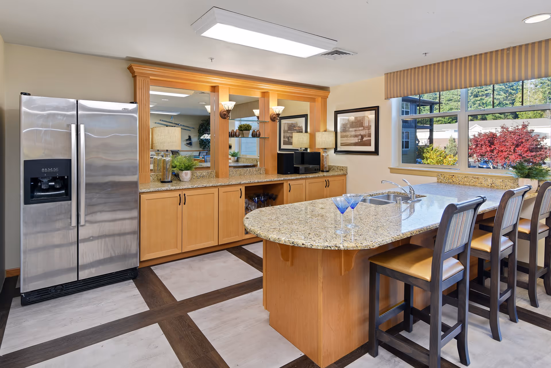 A modern kitchen area with a large granite countertop island featuring a built-in sink and four high chairs. There is a stainless steel refrigerator on the left, wooden cabinets with granite countertops, decorative plants, and framed pictures on the wall. A large window with a striped valance shows a view of trees and buildings outside.