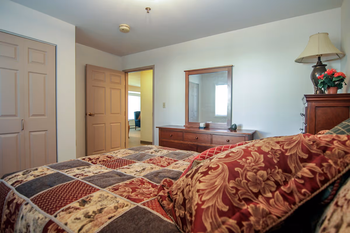 View of a bedroom in Glenwood Senior Apartments featuring a bed with a patterned quilt and pillows, a wooden dresser with a mirror, a lamp, and a potted plant. There are two closed doors and an open doorway leading to another room with a chair visible.
