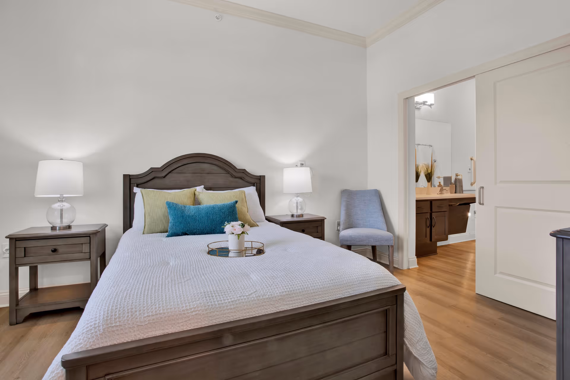 A bedroom with a wooden bed frame and white bedding, decorated with green and blue pillows. There are two matching wooden nightstands with glass lamps on either side of the bed. A small tray with a flower vase is placed on the bed. To the right, there is a blue upholstered chair and an open sliding door leading to a bathroom with a wooden vanity and a mirror.