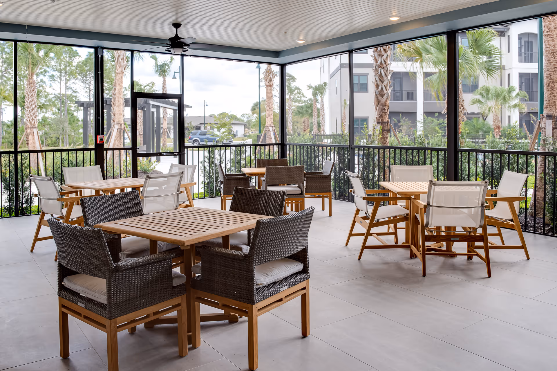 A covered outdoor patio area with several wooden tables and chairs arranged for seating. The patio is enclosed with glass panels and black metal railings, overlooking palm trees and a building in the background.
