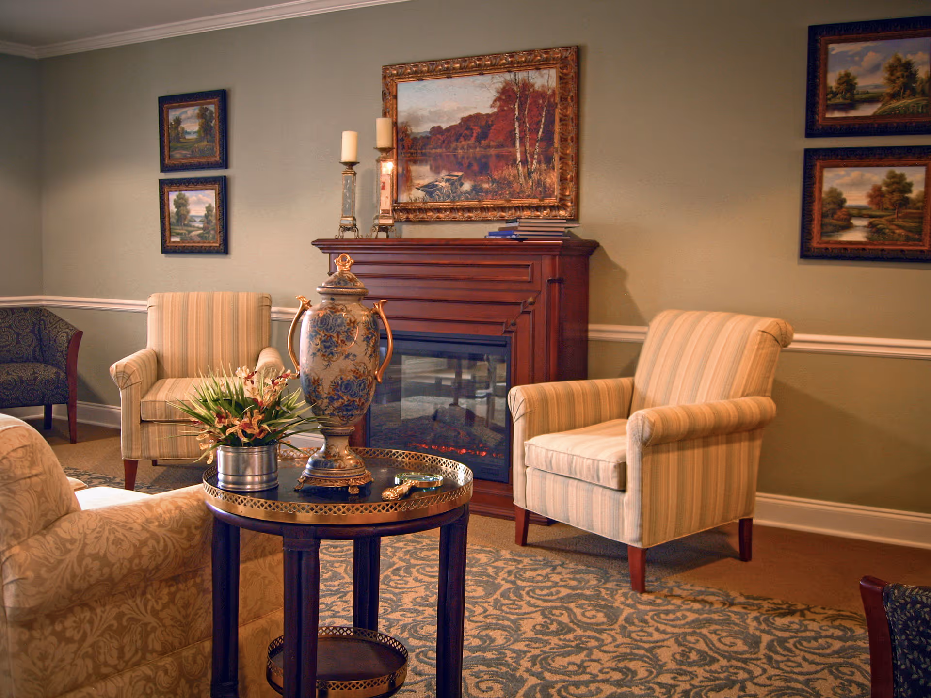 A cozy living room area with two striped armchairs and a patterned armchair arranged around a small round table with a decorative vase and a plant. Behind the chairs is a wooden electric fireplace with a framed landscape painting above it. The walls are adorned with additional framed landscape paintings, and the room has a patterned carpet.