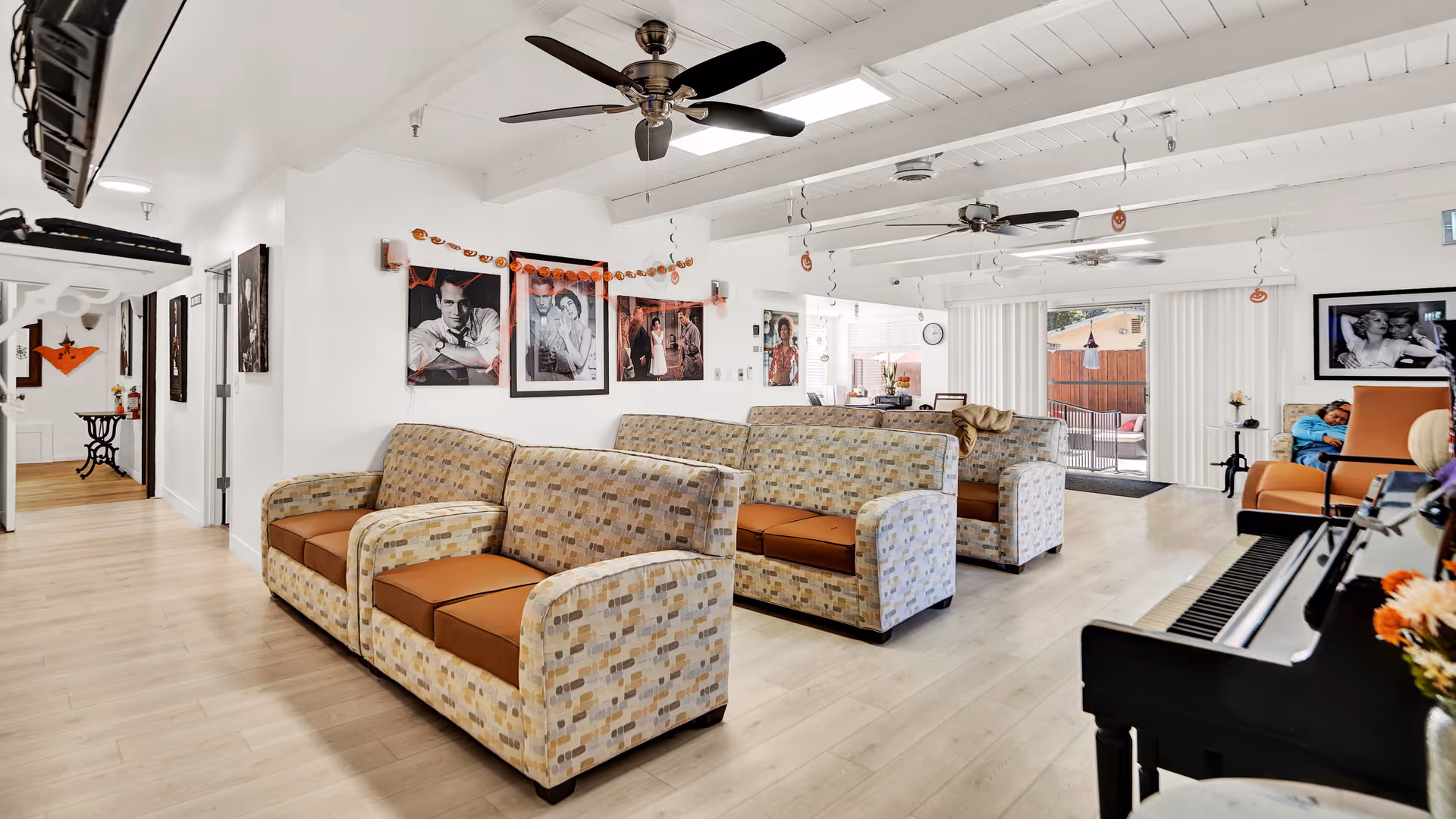 A bright and spacious living room area in a senior living facility with patterned sofas arranged in rows, ceiling fans, and black and white framed photos on the walls. There is a piano on the right side and a person sitting on an orange chair near a sliding glass door leading to an outdoor patio.