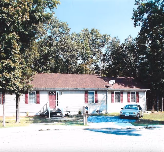 Single-story unit with red shutters, front steps, parked car, mailbox, and trees in yard