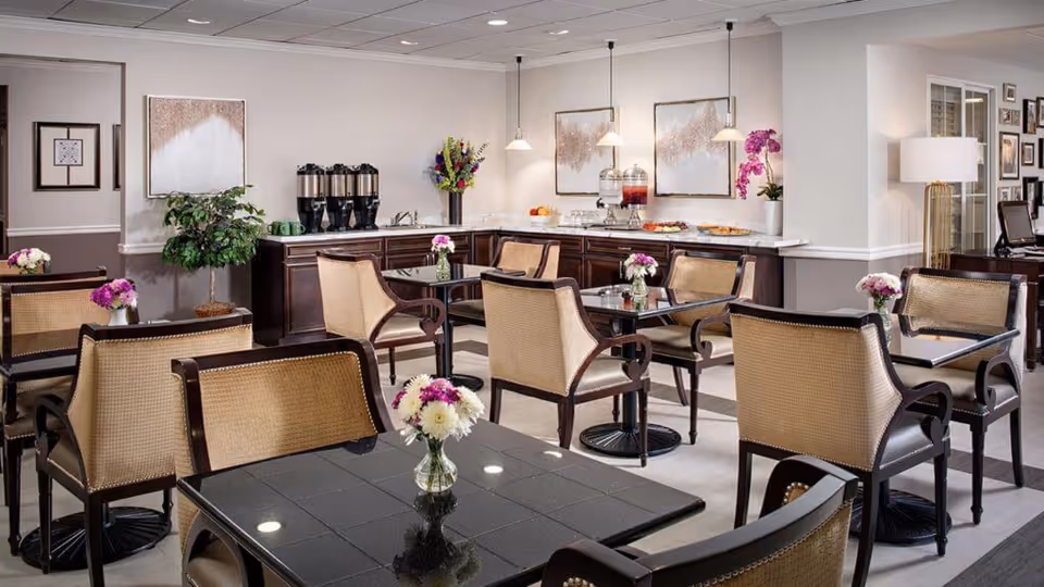 A bright and elegant dining area with multiple tables and cushioned chairs. Each table has a small vase with fresh flowers. In the background, there is a counter with beverage dispensers, fruit bowls, and decorative flowers. The walls are adorned with framed artwork and the space is well-lit with pendant lights and ceiling lights.
