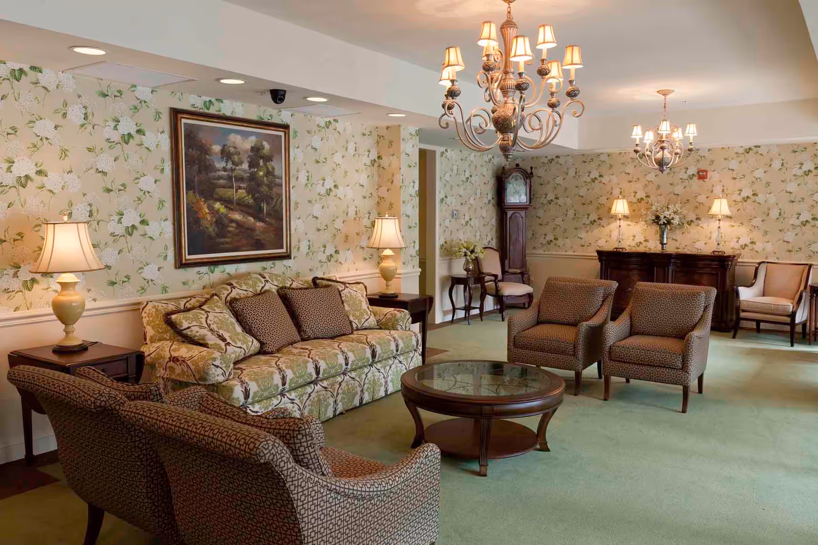A cozy living room with floral wallpaper, a patterned sofa with cushions, two armchairs, a round glass-top coffee table, two chandeliers, table lamps, a grandfather clock, and a wooden sideboard with decorative lamps and flowers.