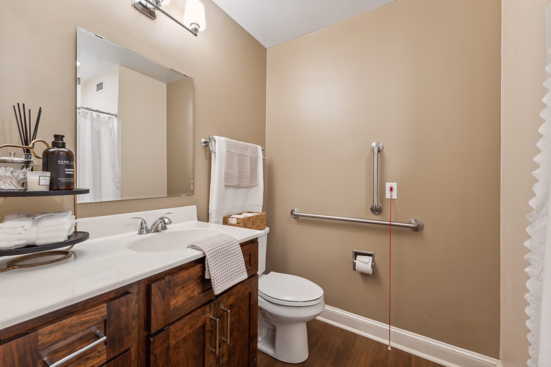 A bathroom with beige walls and wooden floor. It features a white toilet with a grab bar on the wall behind it, a white sink with a wooden cabinet below, a large mirror above the sink, and a towel rack with white towels. There is a red emergency pull cord hanging from a white outlet on the wall near the toilet. A shower curtain is partially visible on the right side.
