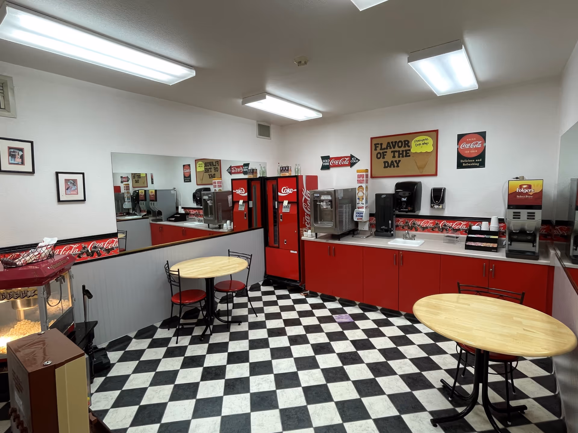 A retro-style snack and beverage area with black and white checkered floor tiles, red cabinets, and Coca-Cola themed decor. There are two small round tables with red cushioned chairs, a popcorn machine, a soda vending machine, coffee dispensers, and a sign on the wall that reads 'FLAVOR OF THE DAY'. The room is brightly lit with fluorescent ceiling lights and has a large mirror on one wall.