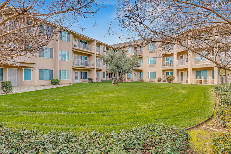 View of a three-story senior living facility building surrounding a well-maintained green lawn with a tree in the center. The building has balconies and large windows, with leafless trees framing the scene under a partly cloudy blue sky.