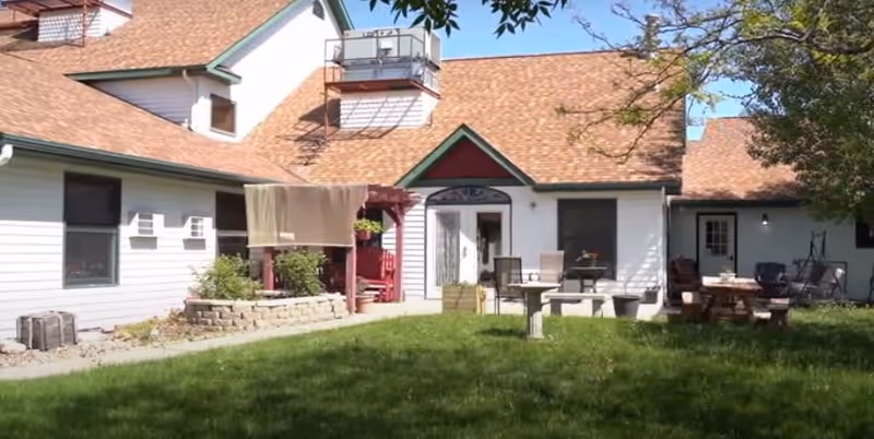 Outdoor view of a residential building with white siding and a brown shingled roof. The scene includes a green lawn, patio furniture, a picnic table, and a tree on the right side. There is a covered porch area with hanging plants and a small stone retaining wall with plants.