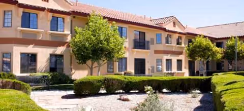 Two-story beige senior living building with red-tiled roof and trimmed shrubs in a sunny courtyard.
