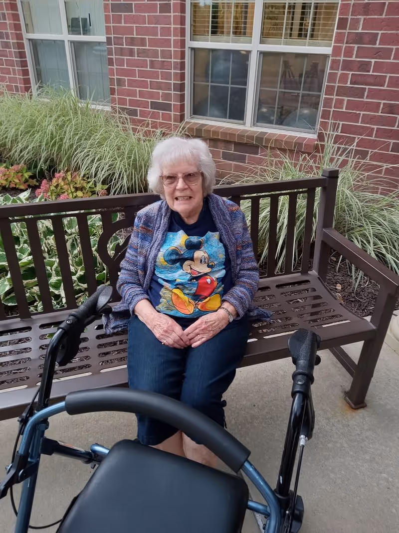 An elderly woman with white hair and glasses sitting on a brown outdoor bench in front of a brick wall with windows and plants. She is wearing a blue Mickey Mouse t-shirt, a patterned cardigan, and dark pants. A walker is positioned in front of her on the concrete ground.