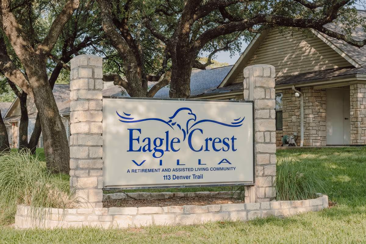 Stone-framed sign in a grassy yard reading "Eagle Crest Villa, a retirement and assisted living community" with a ranch-style building and trees behind it.