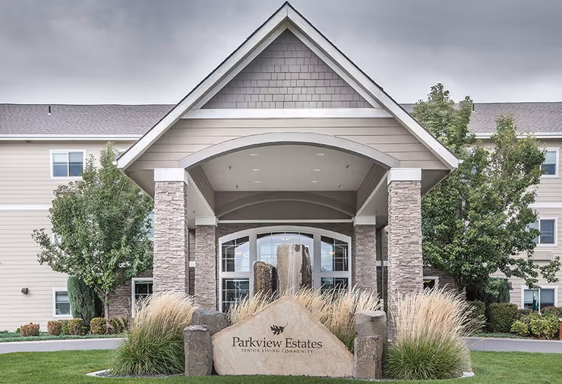 Front exterior view of Parkview Estates senior living community building with a covered entrance supported by stone pillars, surrounded by landscaping including grass, bushes, and trees, and a large stone sign with the facility name in front.