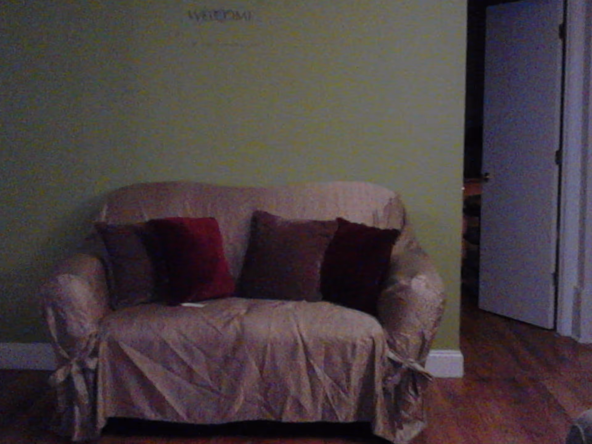 A small loveseat covered with a beige slipcover and adorned with four throw pillows in shades of red and brown, placed against a light green wall with a white baseboard. To the right, there is an open doorway leading to another room with wooden flooring.