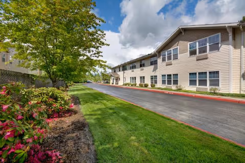 Driveway and landscaped lawn leading to the front exterior of a two-story senior living building under a partly cloudy sky.