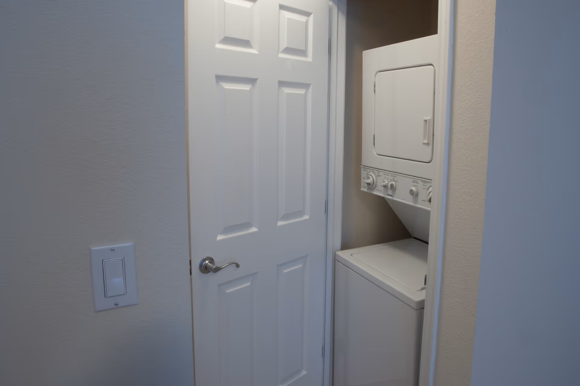 A small laundry closet with a stacked white washer and dryer unit partially visible behind a white door with a silver handle. A light switch is mounted on the wall to the left of the door.