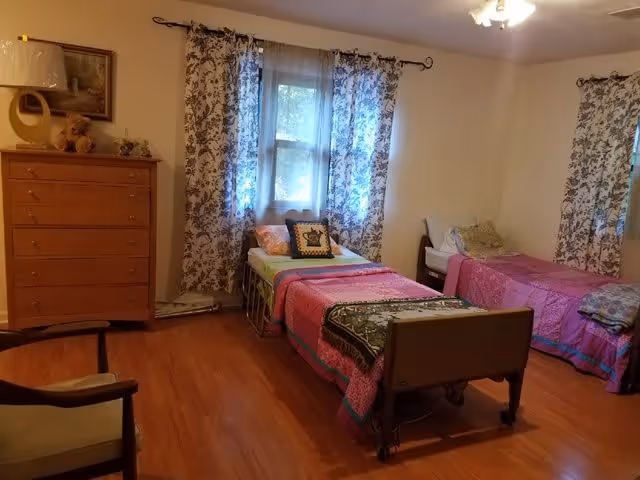 A residential care home bedroom with two single beds covered in colorful quilts. Each bed has pillows and is positioned against the walls with windows behind them, adorned with floral curtains. There is a wooden dresser with decorative items on top and a framed picture hanging above it. A wooden chair with armrests is partially visible in the foreground. The room has wooden flooring and neutral-colored walls.