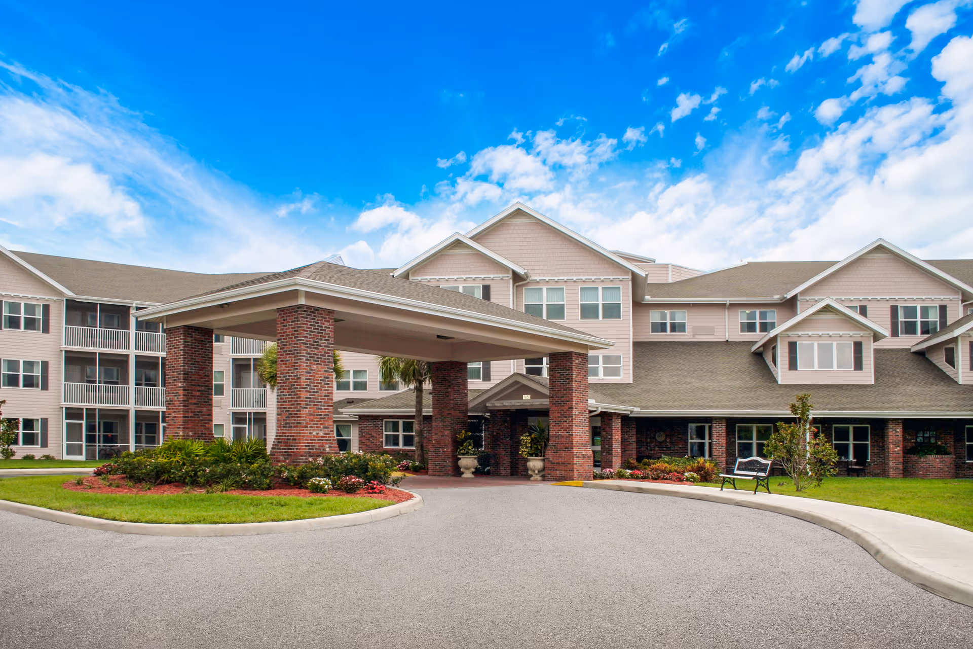 Front entrance of a multi-story senior living building with a covered porte-cochère, landscaped driveway and bench.
