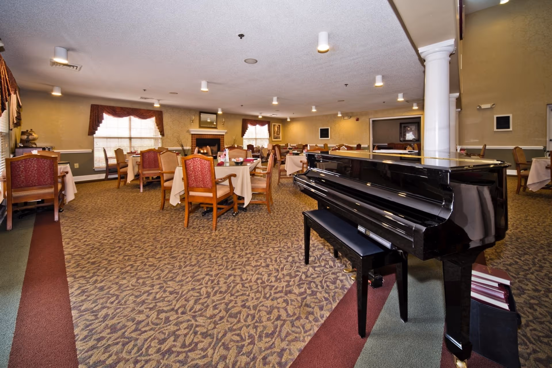 A spacious dining room with multiple tables covered in beige tablecloths and surrounded by wooden chairs with red and beige upholstery. A black grand piano with a matching bench is positioned in the foreground on the right side. The room features patterned carpet, large windows with red valances, a fireplace, and soft ceiling lighting.