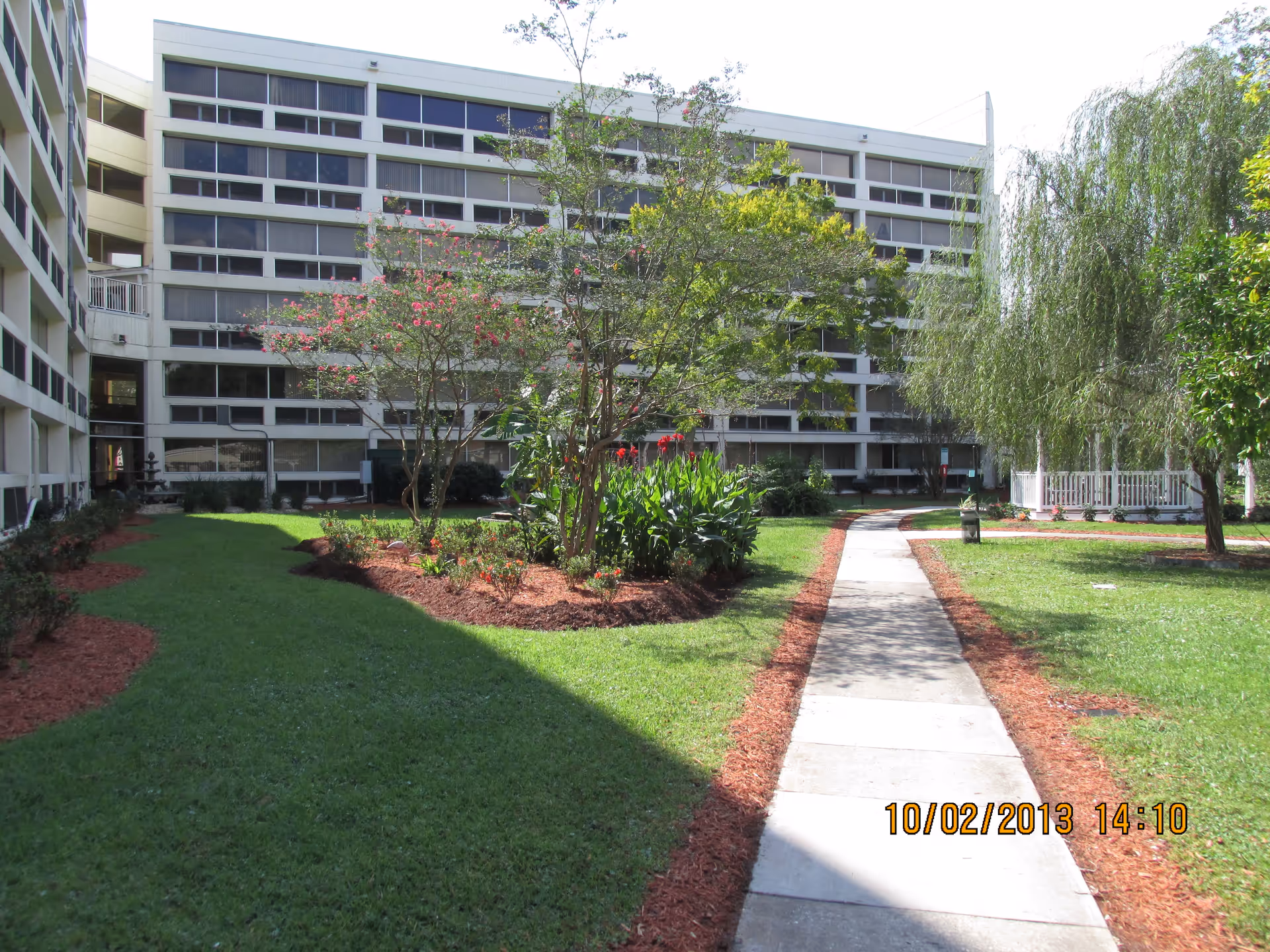 Outdoor view of a senior living facility with a concrete walkway leading through a landscaped garden area with green grass, flowering plants, and trees. In the background, there is a multi-story building with many windows and balconies. The sky is clear and the scene is well-lit by daylight.
