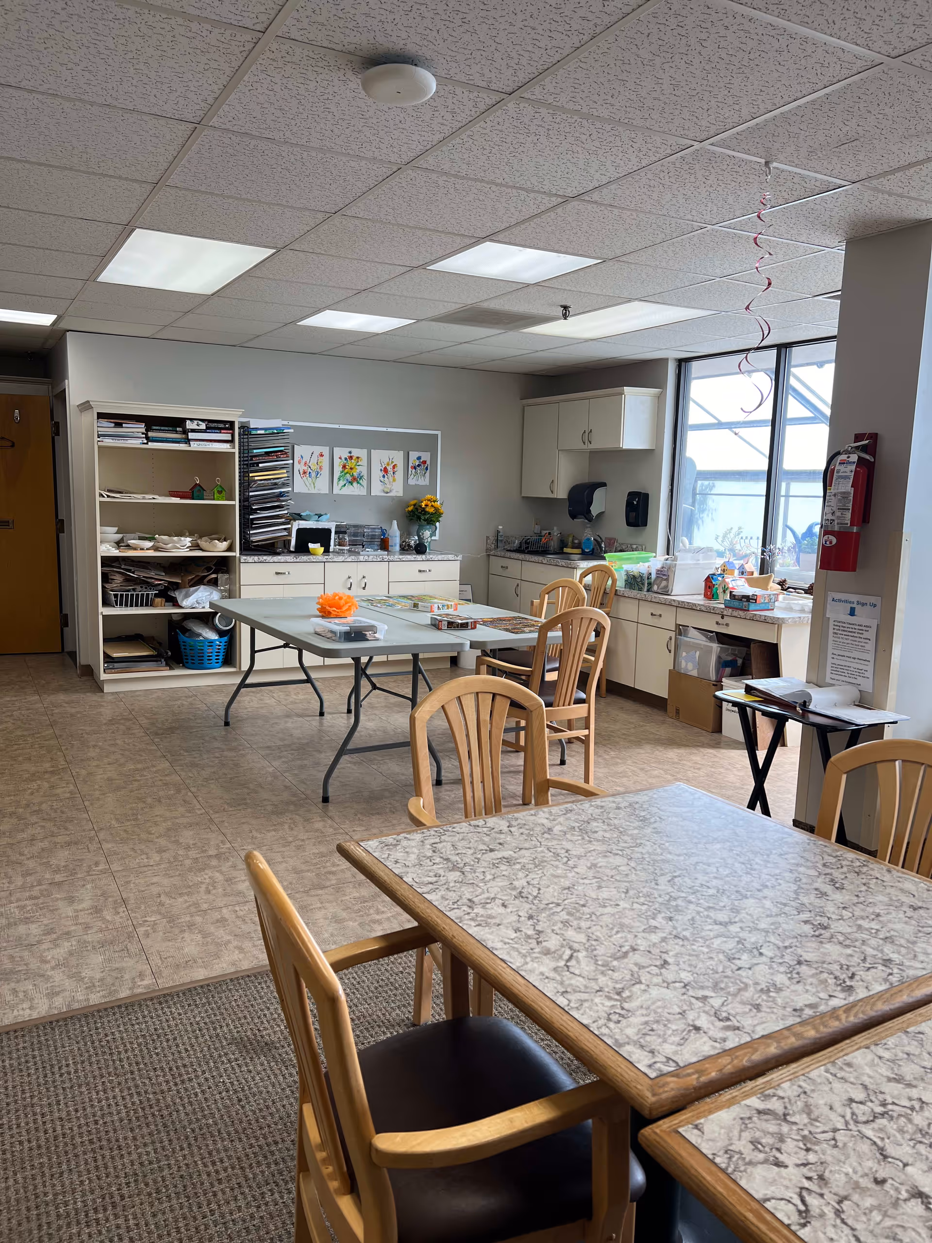 A well-lit activity room with tables and wooden chairs. The room has a tiled floor and a drop ceiling with fluorescent lights. There are cabinets along the back wall with art supplies and colorful paintings displayed above. A large window lets in natural light, and a fire extinguisher is mounted on a pillar near the window. A sign-up sheet for activities is visible on a small table near the pillar.