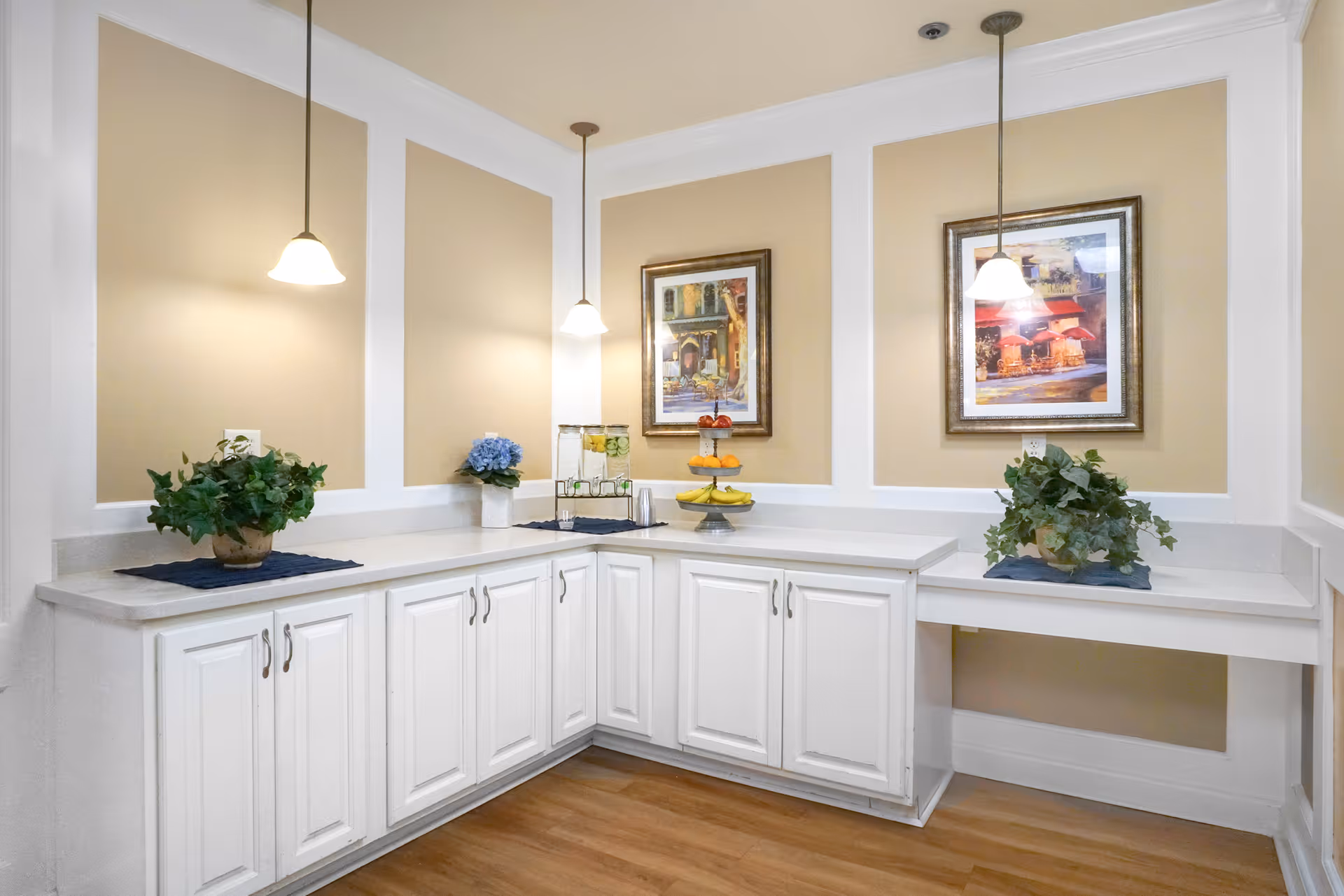 A bright corner countertop area with white cabinets and beige walls, decorated with two framed paintings, two hanging pendant lights, two potted plants, a water dispenser, and a tiered fruit stand with bananas and oranges.