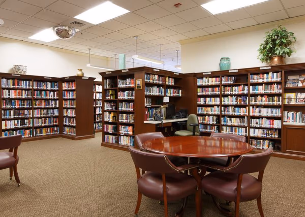 Well-lit interior library with wooden bookshelves, a round polished table and chairs, and a small computer station.