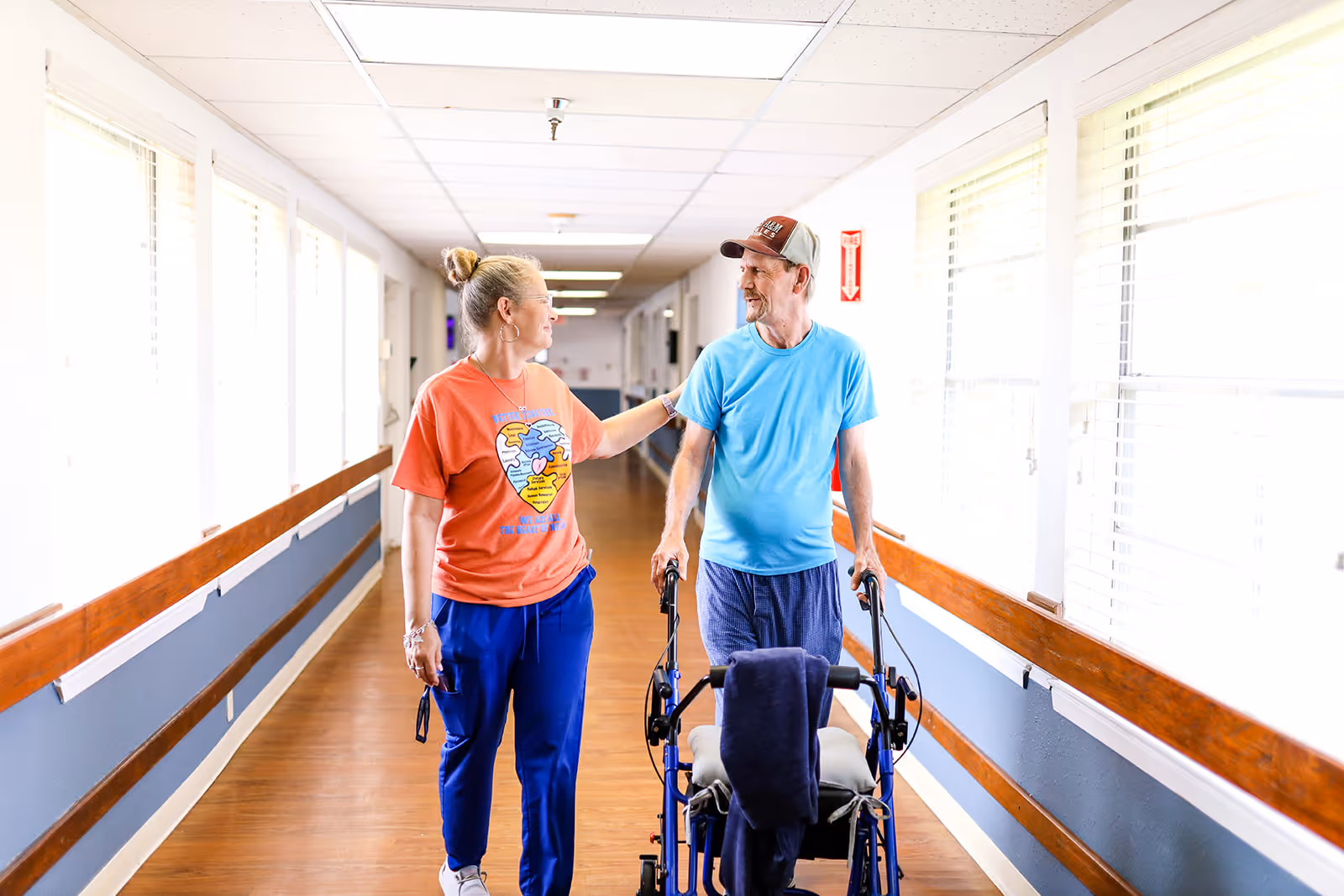 A caregiver in an orange shirt and blue pants walks alongside an elderly man using a walker in a brightly lit hallway with wooden handrails and large windows.