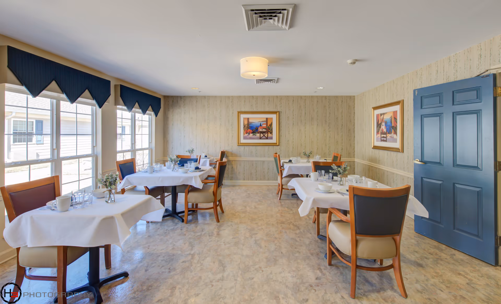 A small dining room with four tables covered with white tablecloths, each set with cups, bowls, glasses, and small vases with greenery. The room has large windows with blue valances on the left side, beige patterned wallpaper, two framed paintings on the walls, and a blue door on the right side. The floor is a light-colored tile.
