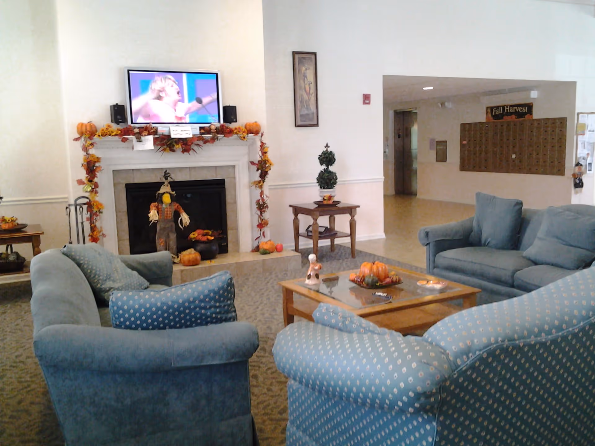 A cozy senior living facility common area with blue upholstered sofas and armchairs arranged around a wooden coffee table. The fireplace mantel is decorated with autumn-themed garlands, small pumpkins, and a scarecrow figure. A flat-screen TV is mounted above the fireplace. In the background, there is a hallway with mailboxes and a bulletin board.