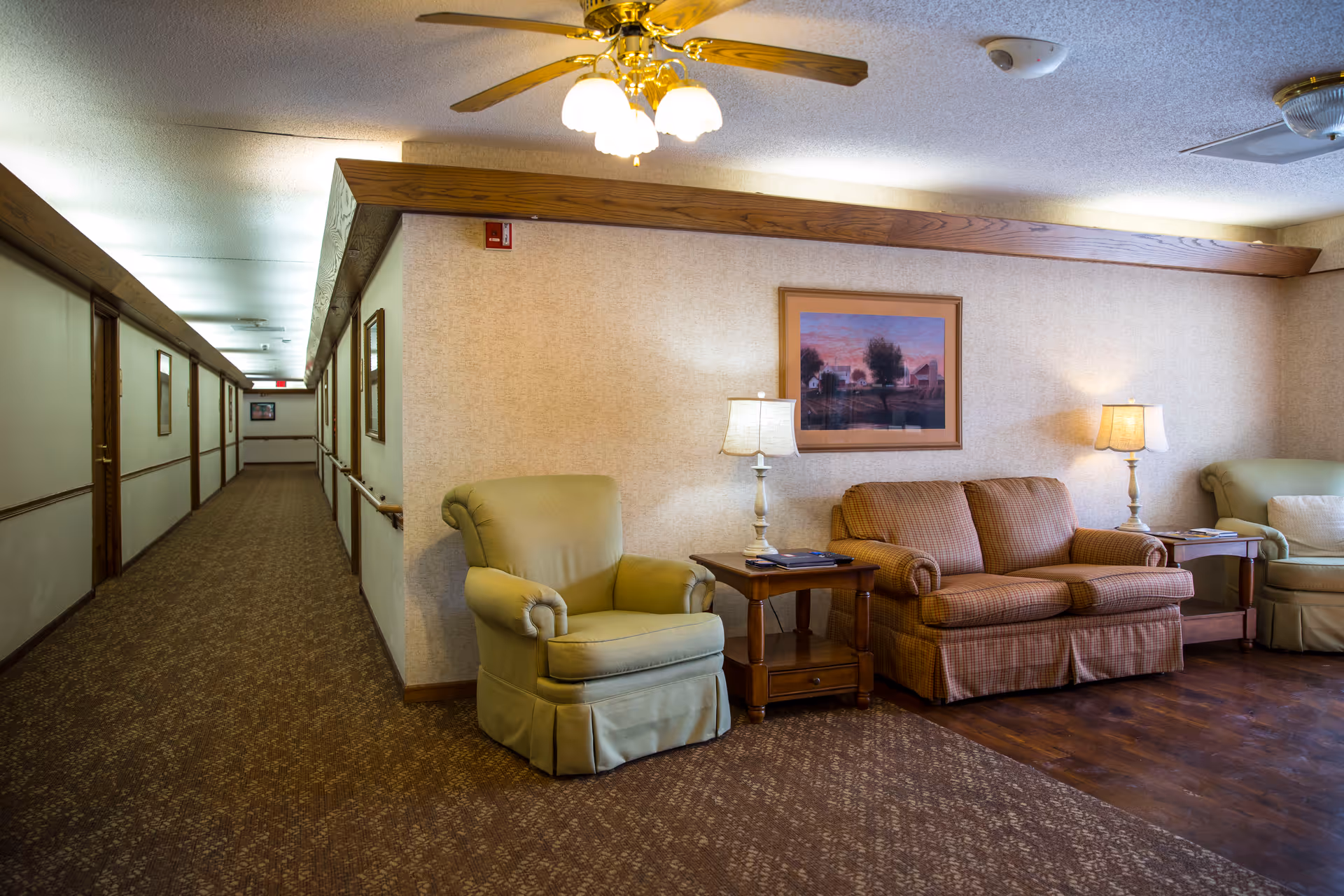 A cozy sitting area in a senior living facility with two armchairs and a loveseat arranged around two wooden side tables with lamps. A framed picture hangs on the wall above the loveseat. To the left, a long hallway with closed doors and handrails extends into the distance. The ceiling has a ceiling fan with lights.