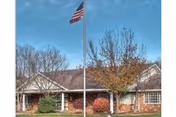 Front exterior of a single-story brick senior living building with an American flag on a flagpole, shrubs, and trees in front.