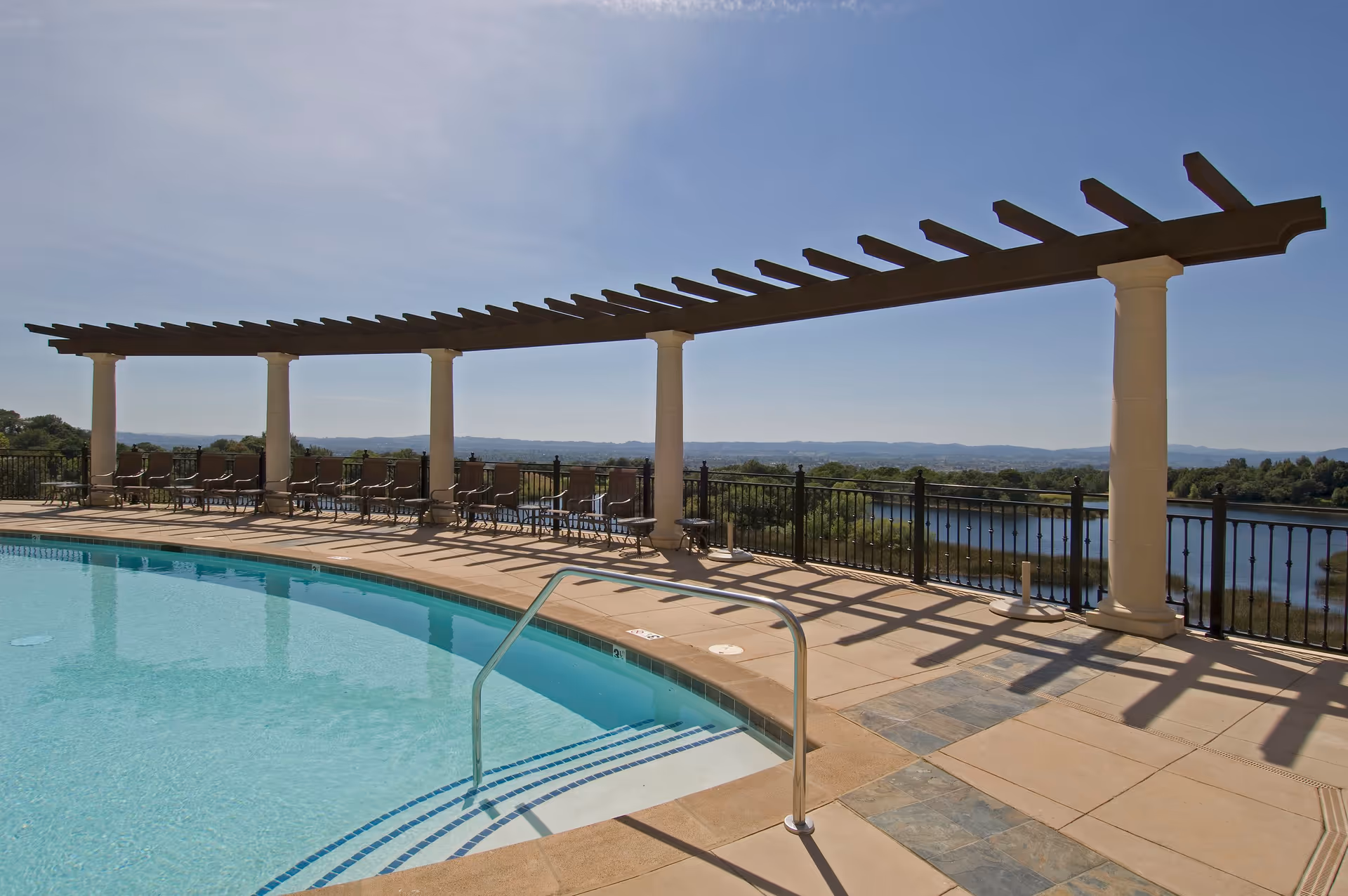 Rooftop swimming pool and deck with a pergola, lounge chairs and a railing overlooking a lake and distant hills.