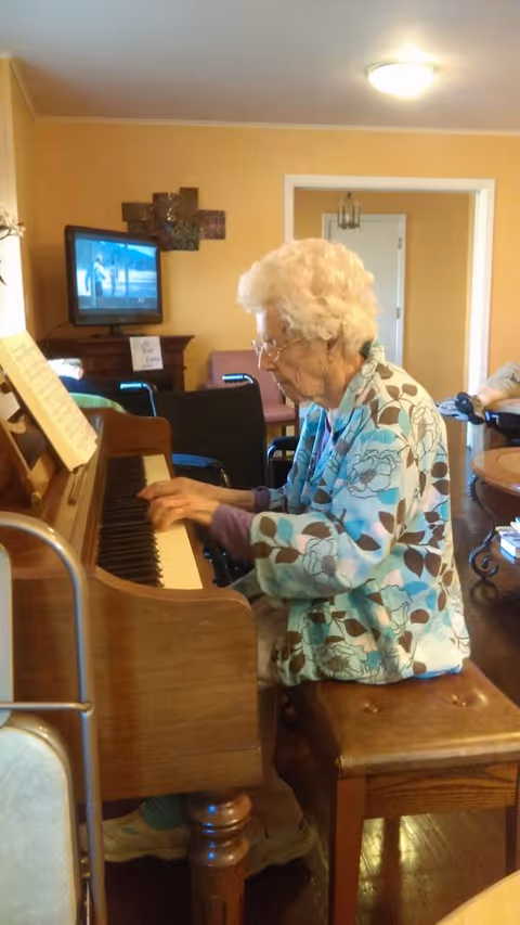 An elderly woman with white hair and glasses is sitting on a wooden bench playing an upright piano in a cozy room with yellow walls. A television is mounted on the wall in the background, and there are chairs and a table visible in the room.