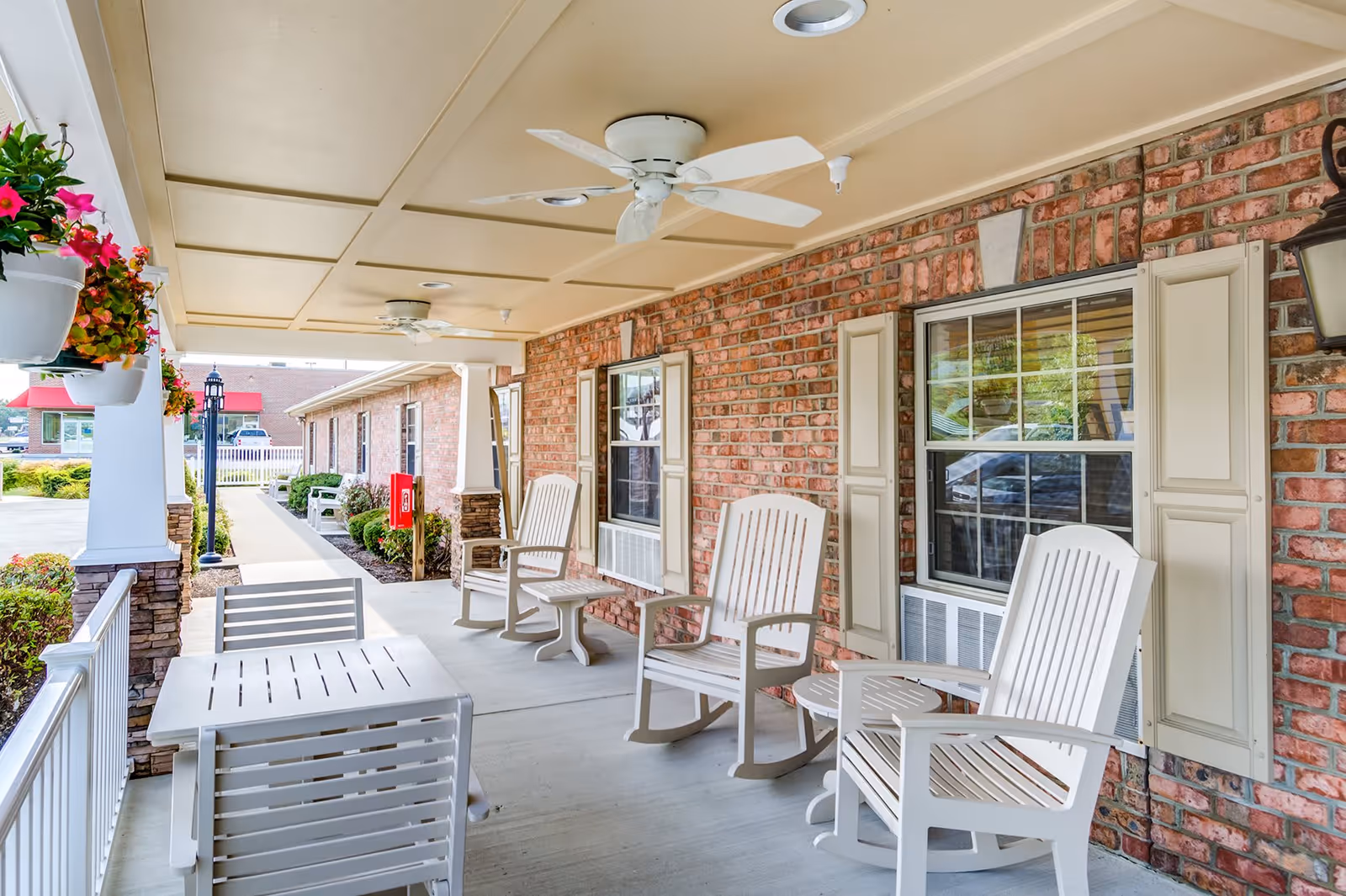 Covered outdoor patio area with white rocking chairs and tables, ceiling fans, hanging flower pots, and a brick wall with windows and shutters.