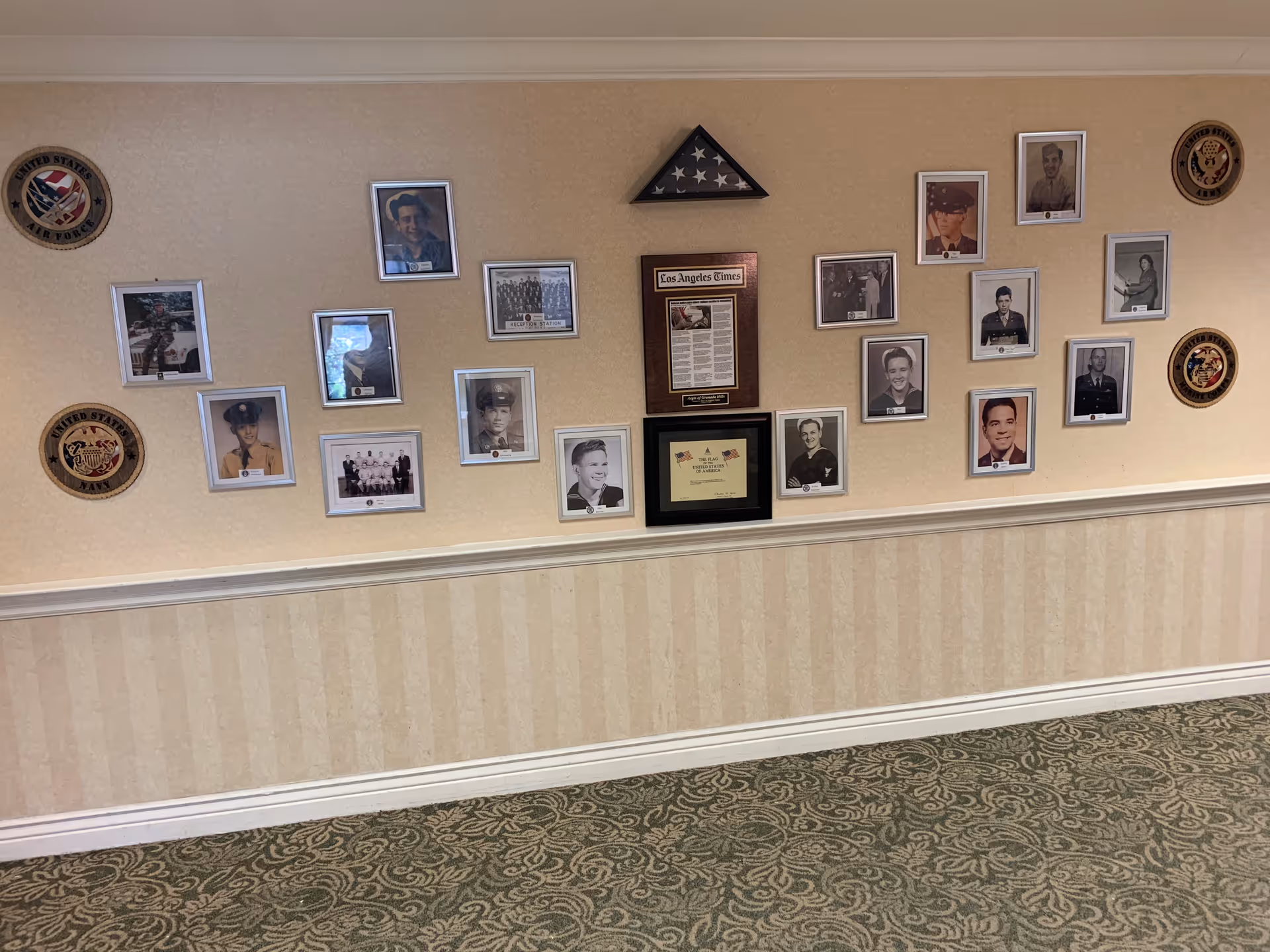 A wall display featuring framed photographs of military personnel, plaques, and military insignia emblems from different branches of the United States Armed Forces. The wall has a beige wallpaper with a white chair rail and patterned carpet below.