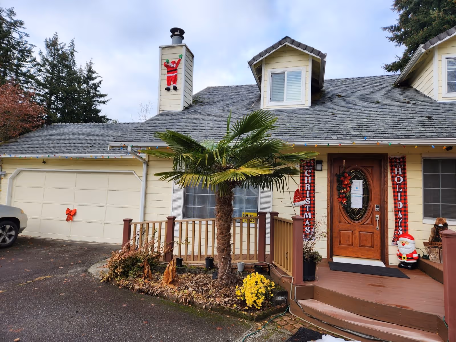 Front exterior view of a yellow house with a gray roof decorated for Christmas. There is a Santa Claus figure climbing the chimney, Christmas lights along the roof edge, and red banners on either side of the wooden front door that read 'Christmas' and 'Holidays'. A small palm tree and some plants are in front of the porch, and a small Santa decoration is near the door. A car is partially visible on the left side in the driveway.