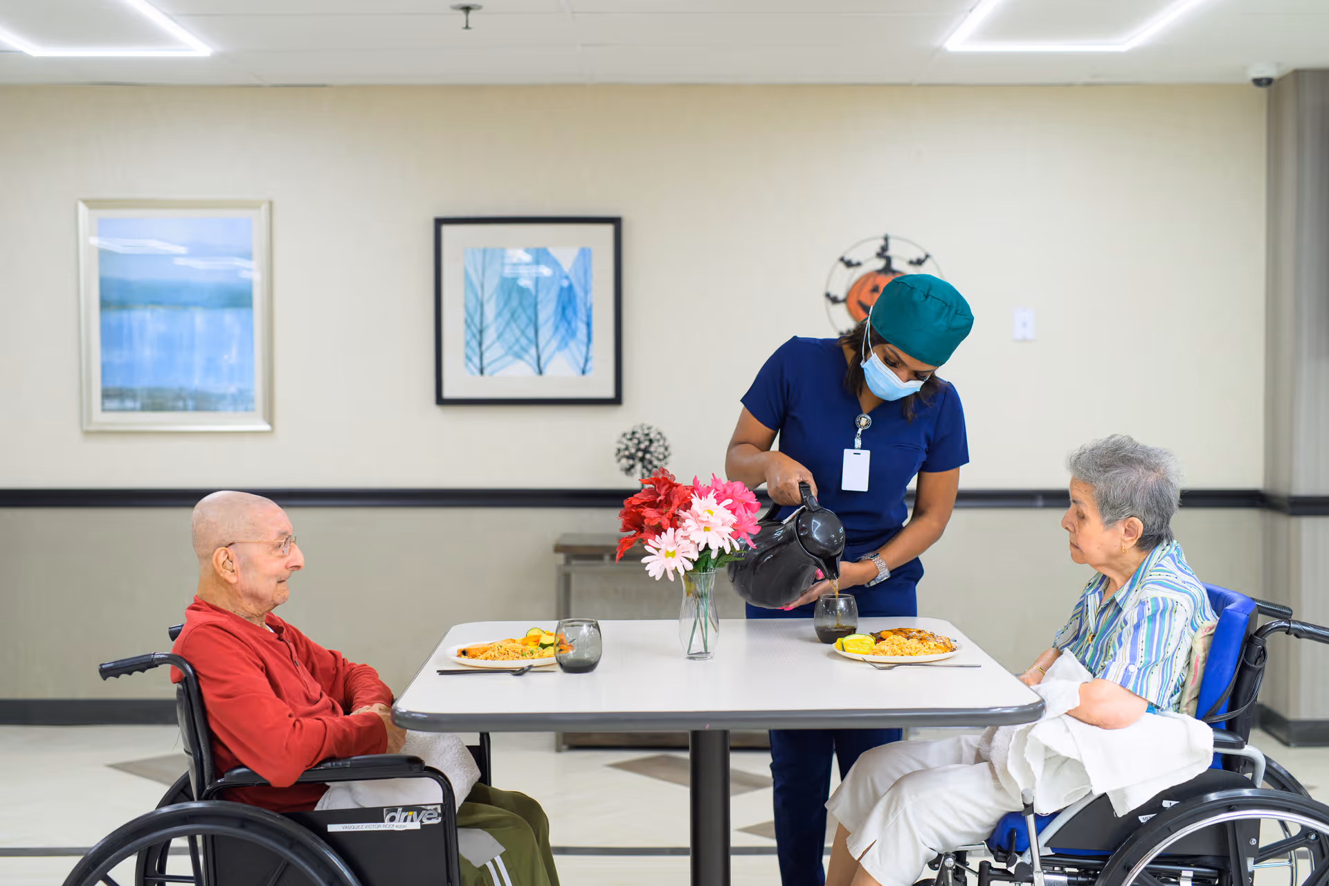 A healthcare worker wearing a mask and scrubs is pouring a drink from a black pitcher into a glass for an elderly woman seated in a wheelchair at a dining table. Another elderly man in a wheelchair sits across from her. Both have plates of food in front of them. The room has light-colored walls with framed artwork and a flower vase on the table.
