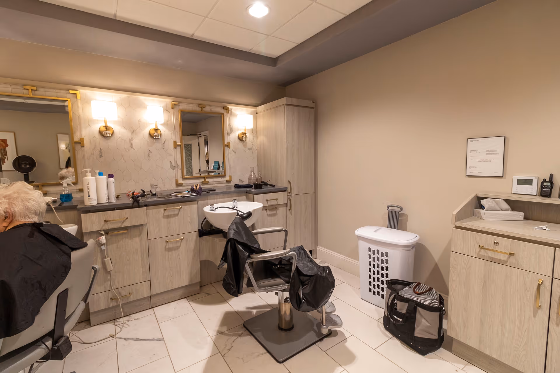 Interior of a hair salon area in a senior living facility with a styling chair covered with a black cape, a washing station, mirrors with wall-mounted lights, cabinets, and a laundry basket. An elderly person is seated in one of the chairs.