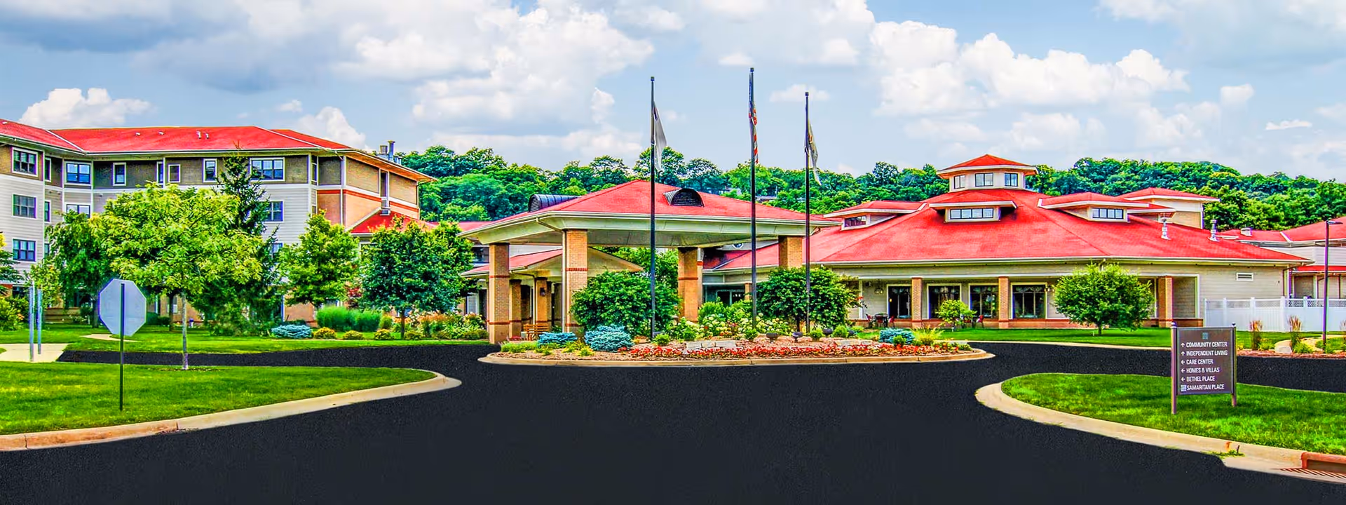 Front exterior view of EverTrue Lutheran Hillside Village showing a large building with red roofs, multiple windows, and a covered entrance. The surrounding area includes well-maintained green lawns, trees, and a circular driveway with flagpoles in the center.